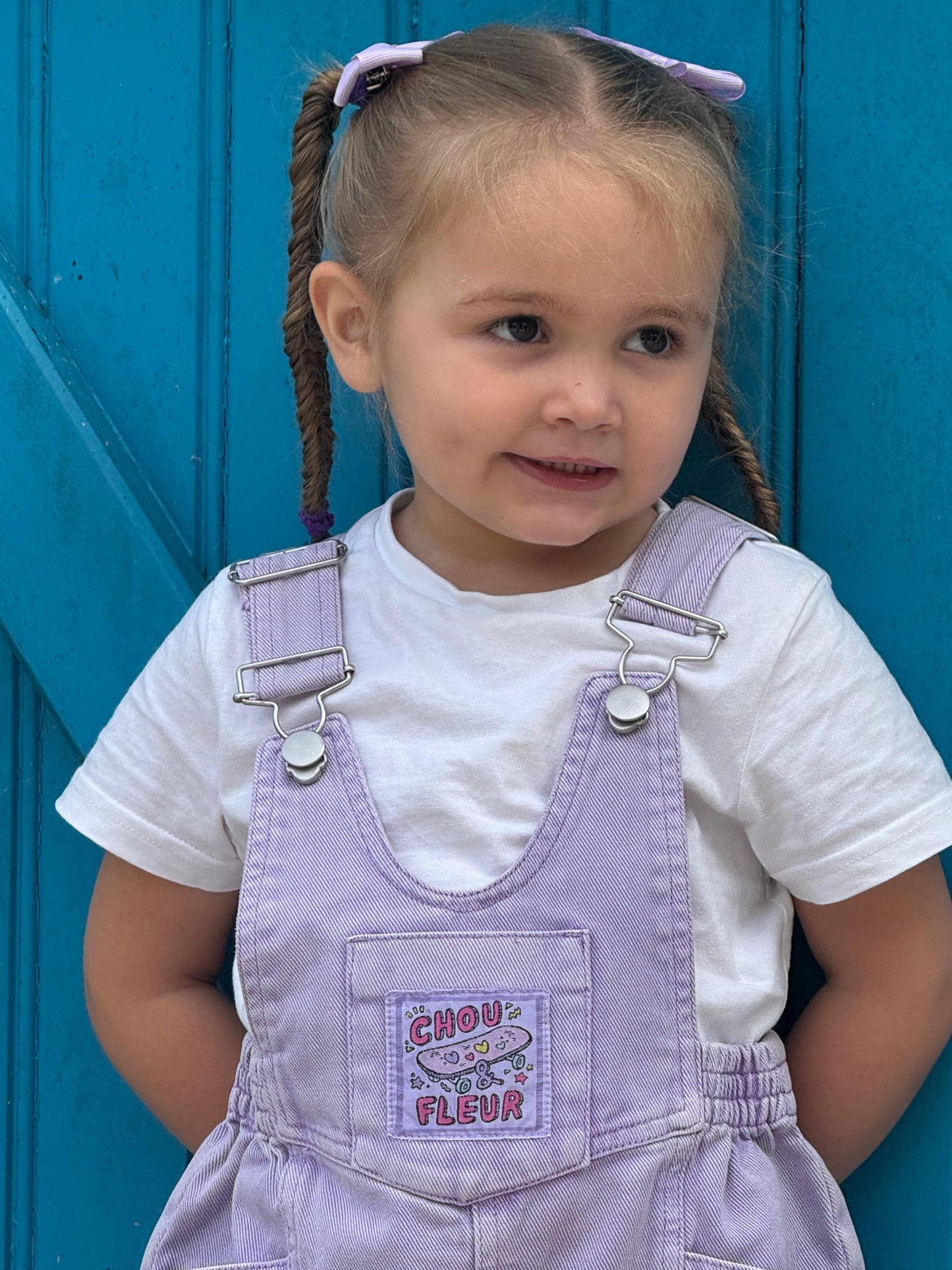 Fille 3 ans en salopette mauve les mains dans le dos