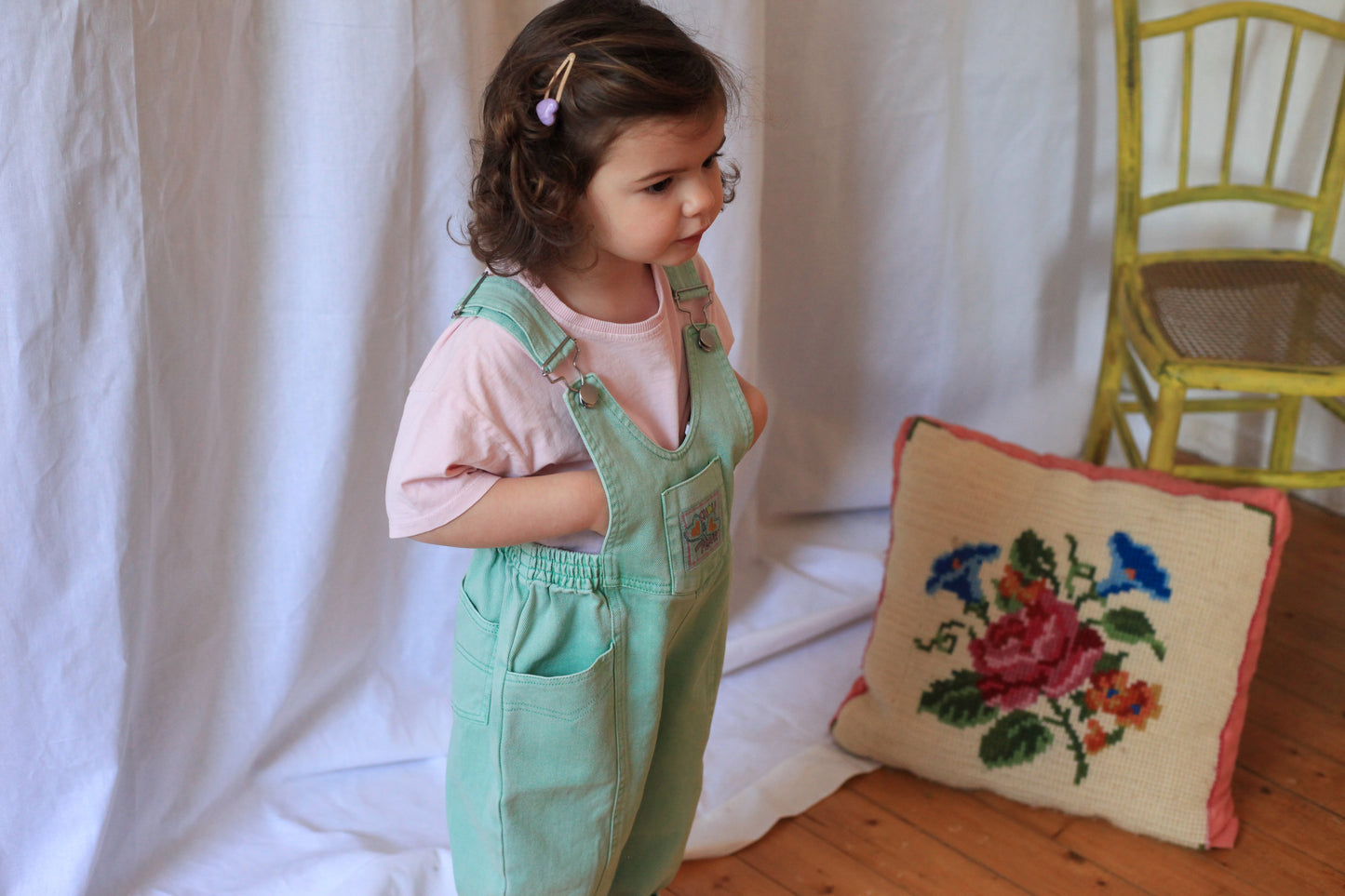 Petite fille de profil en salopette verte vintage et t-shirt rose, debout près d'un coussin brodé floral et d'une chaise jaune