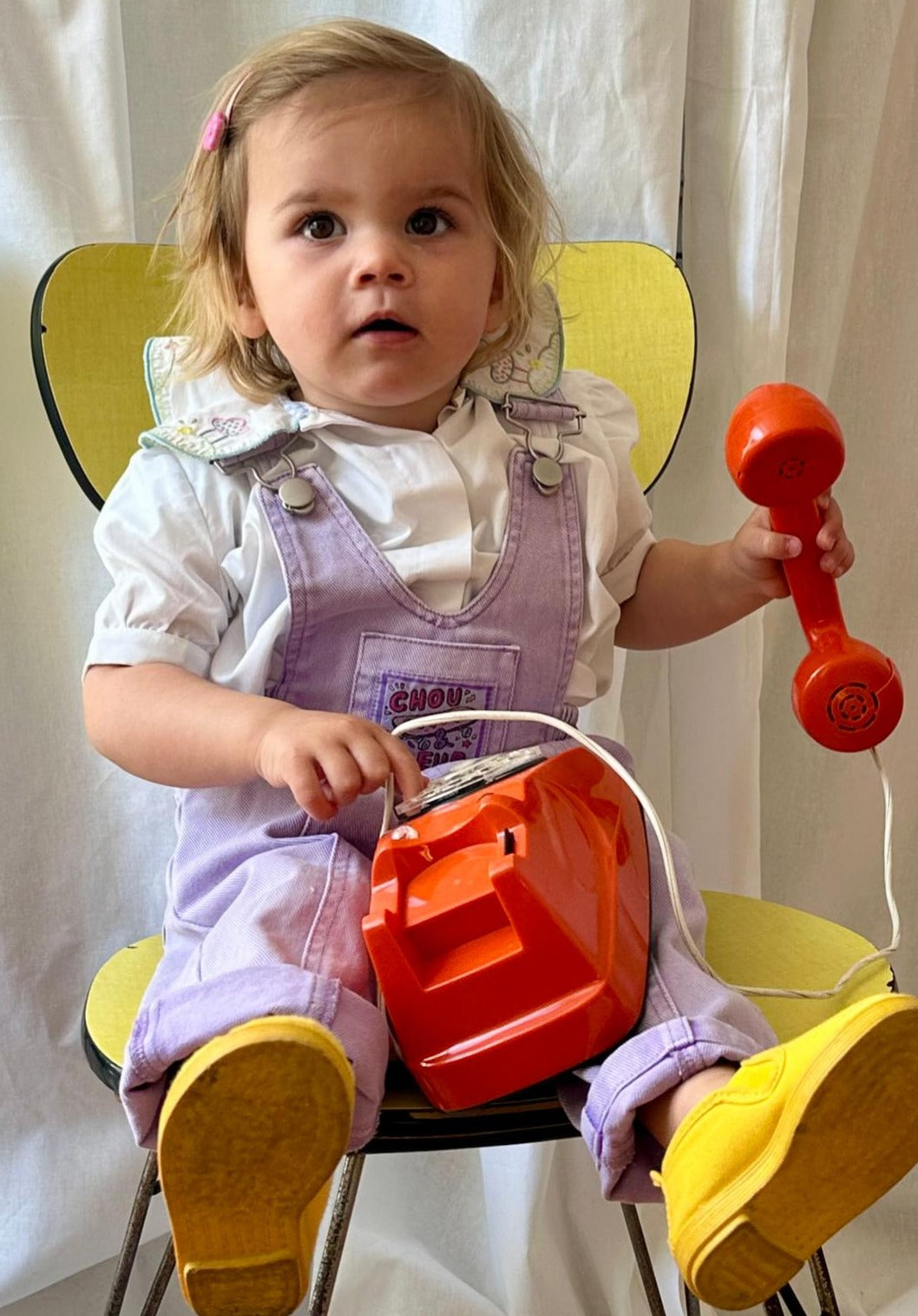Bébé assis en salopette mauve vintage tenant un téléphone jouet orange, sur une chaise jaune rétro