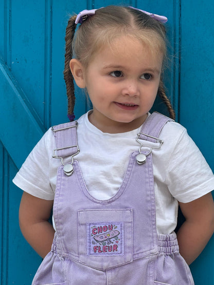 Fille 3 ans en salopette mauve les mains dans le dos