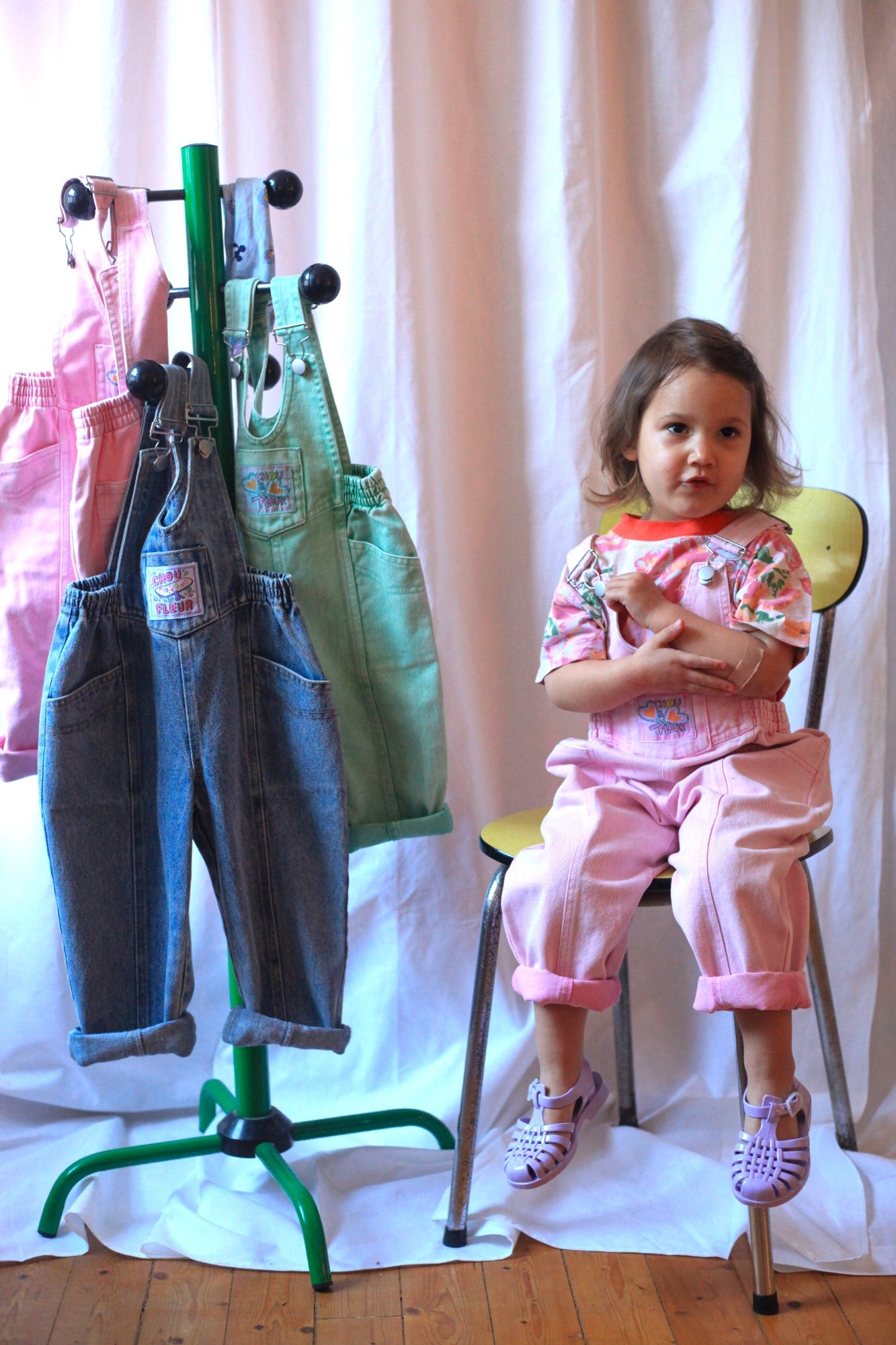 Petite fille assise en salopette rose vintage avec t-shirt fleuri, devant un portant de salopettes colorées pour enfants