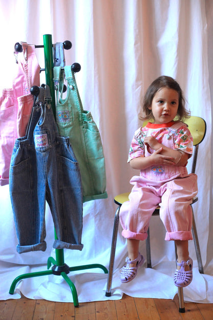 Petite fille assise en salopette rose vintage avec t-shirt fleuri, devant un portant de salopettes colorées pour enfants