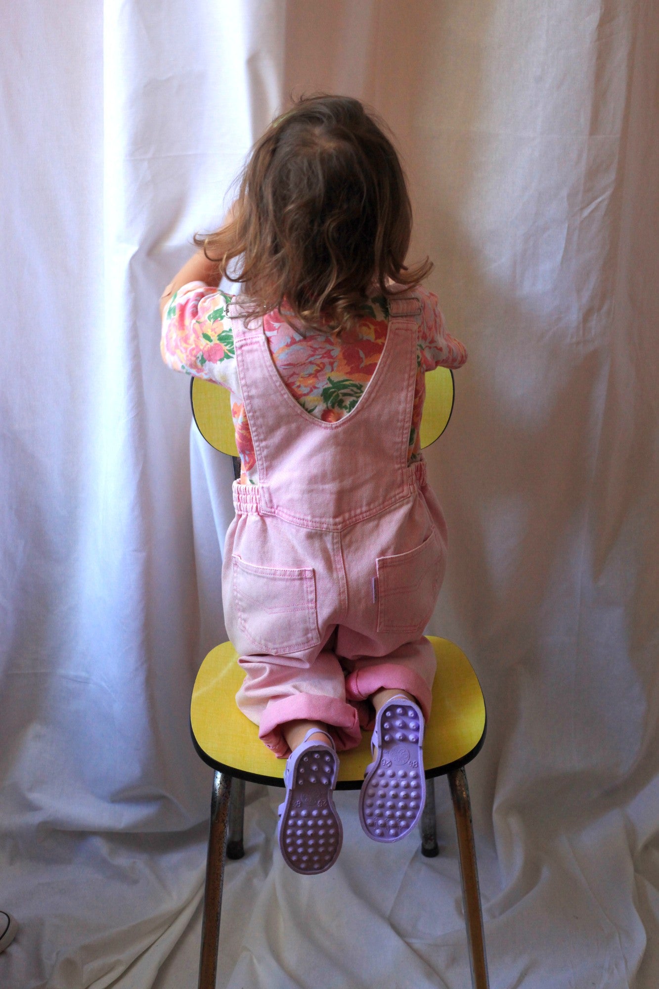 Vue de dos d'une enfant en salopette rose vintage debout sur une chaise, t-shirt fleuri visible sous les bretelles croisées
