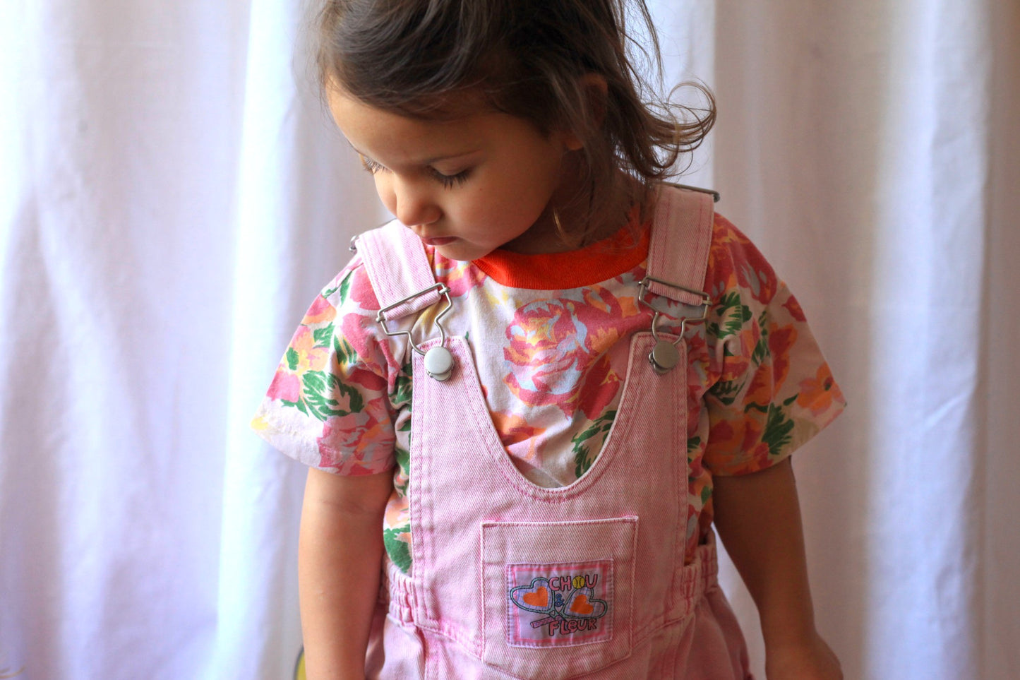 Fillette regardant vers le bas, portant une salopette rose vintage avec un t-shirt fleuri à dominante rose et orange