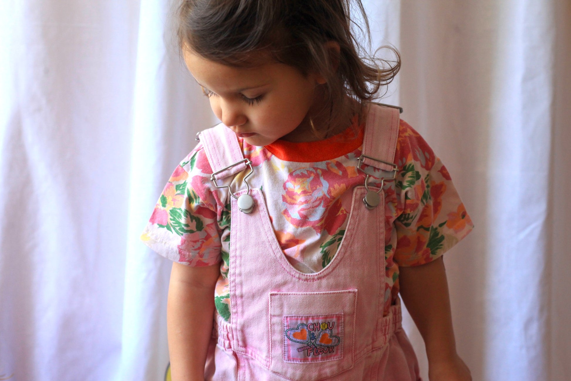 Fillette regardant vers le bas, portant une salopette rose vintage avec un t-shirt fleuri à dominante rose et orange