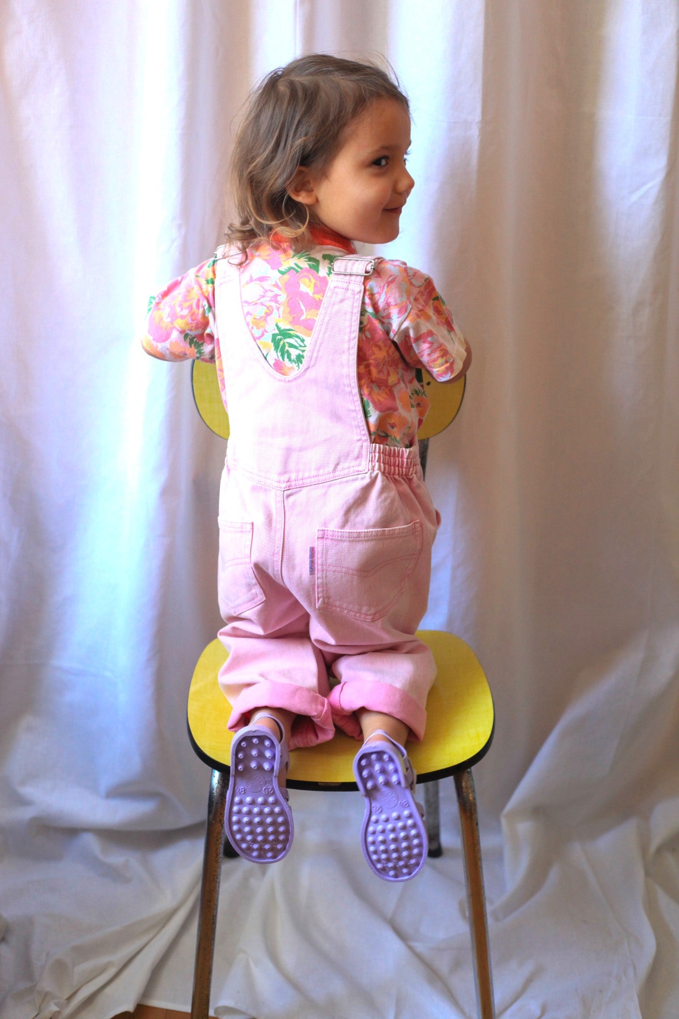 Fillette souriante de dos en salopette rose vintage et t-shirt fleuri, debout sur une chaise jaune rétro
