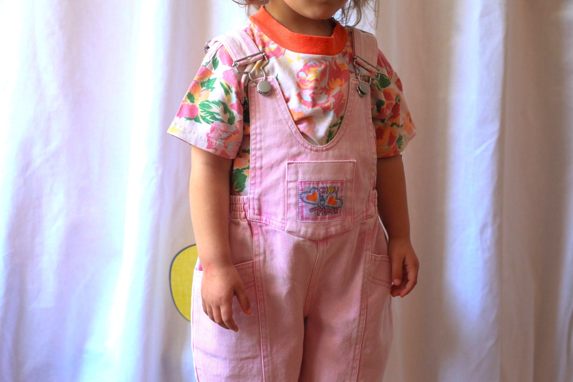 Vue partielle d'une enfant en salopette rose vintage avec t-shirt fleuri, debout devant un fond clair