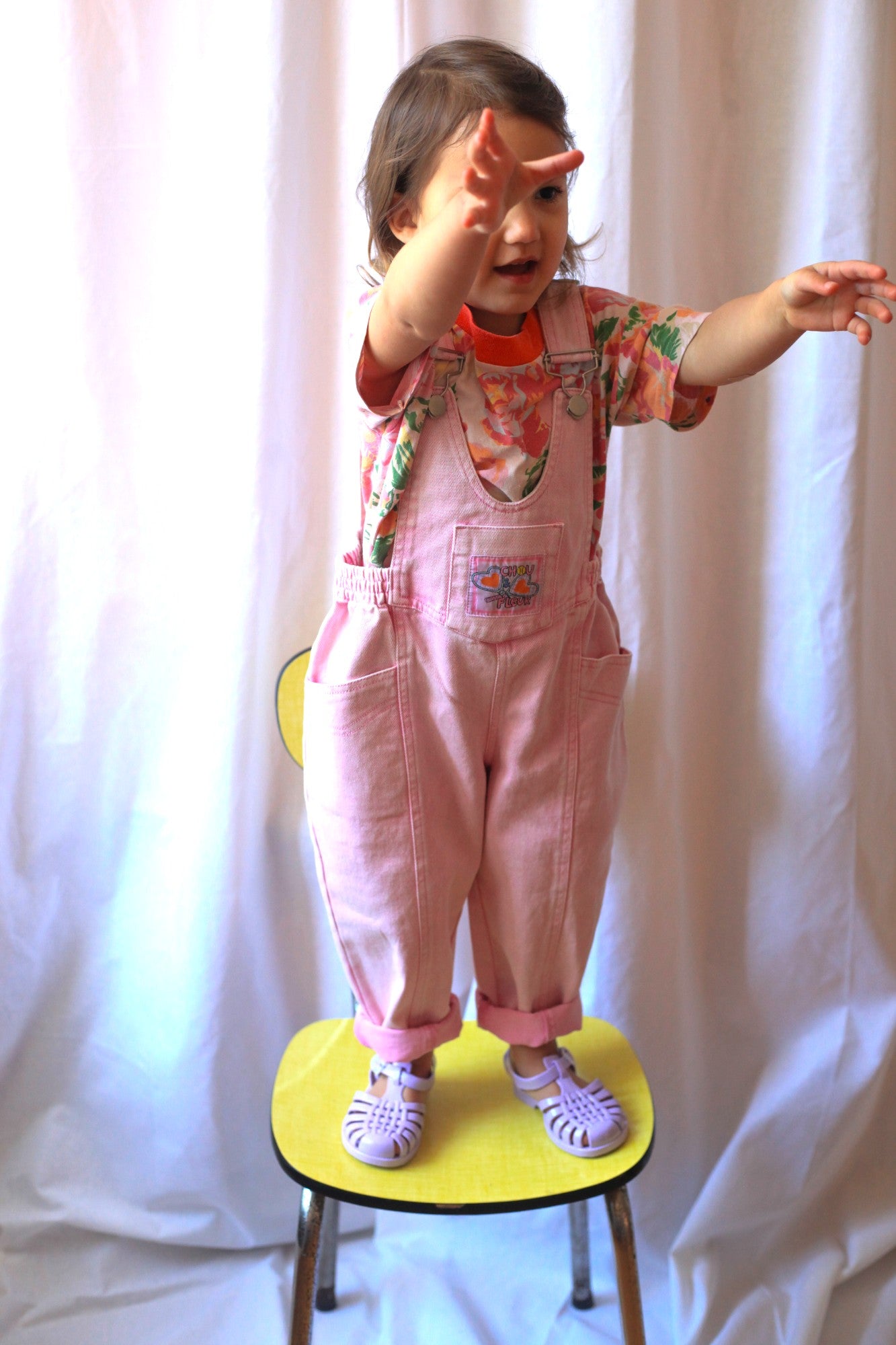 Petite fille en salopette rose vintage et t-shirt fleuri, bras tendus debout sur une chaise jaune devant un rideau blanc