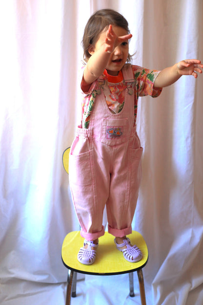 Petite fille en salopette rose vintage et t-shirt fleuri, bras tendus debout sur une chaise jaune devant un rideau blanc