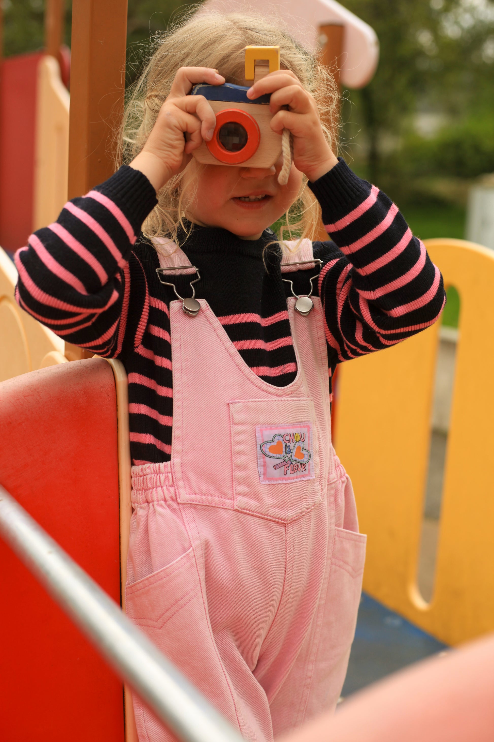 Enfant en salopette rose vintage avec marinière noir et rose, jouant avec un appareil photo jouet dans une aire de jeu