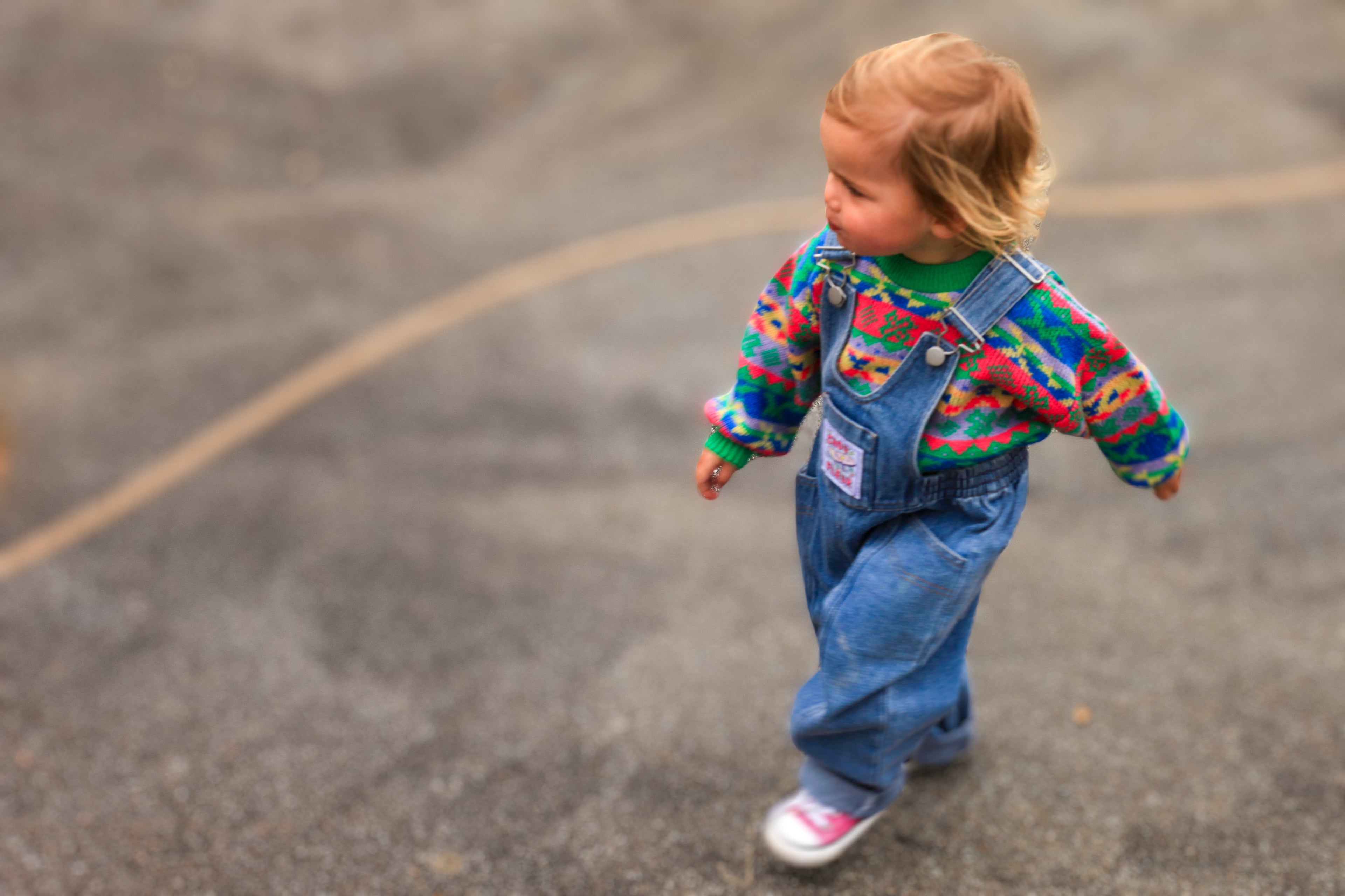 Jeune enfant en salopette vintage en denim bleu et pull coloré, marchant à l'extérieur sur un sol en bitume